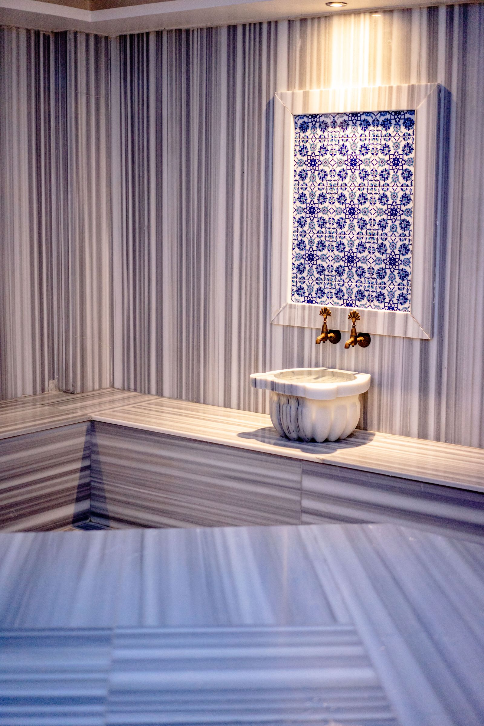 Inside the Moroccan Hammam Ritual at Kimantra: A Step-by-Step Photo Story 3 Traditional marble hammam interior with striped marble walls, a heated bench, and a white carved kurna basin beneath a blue Iznik-tile panel with brass taps