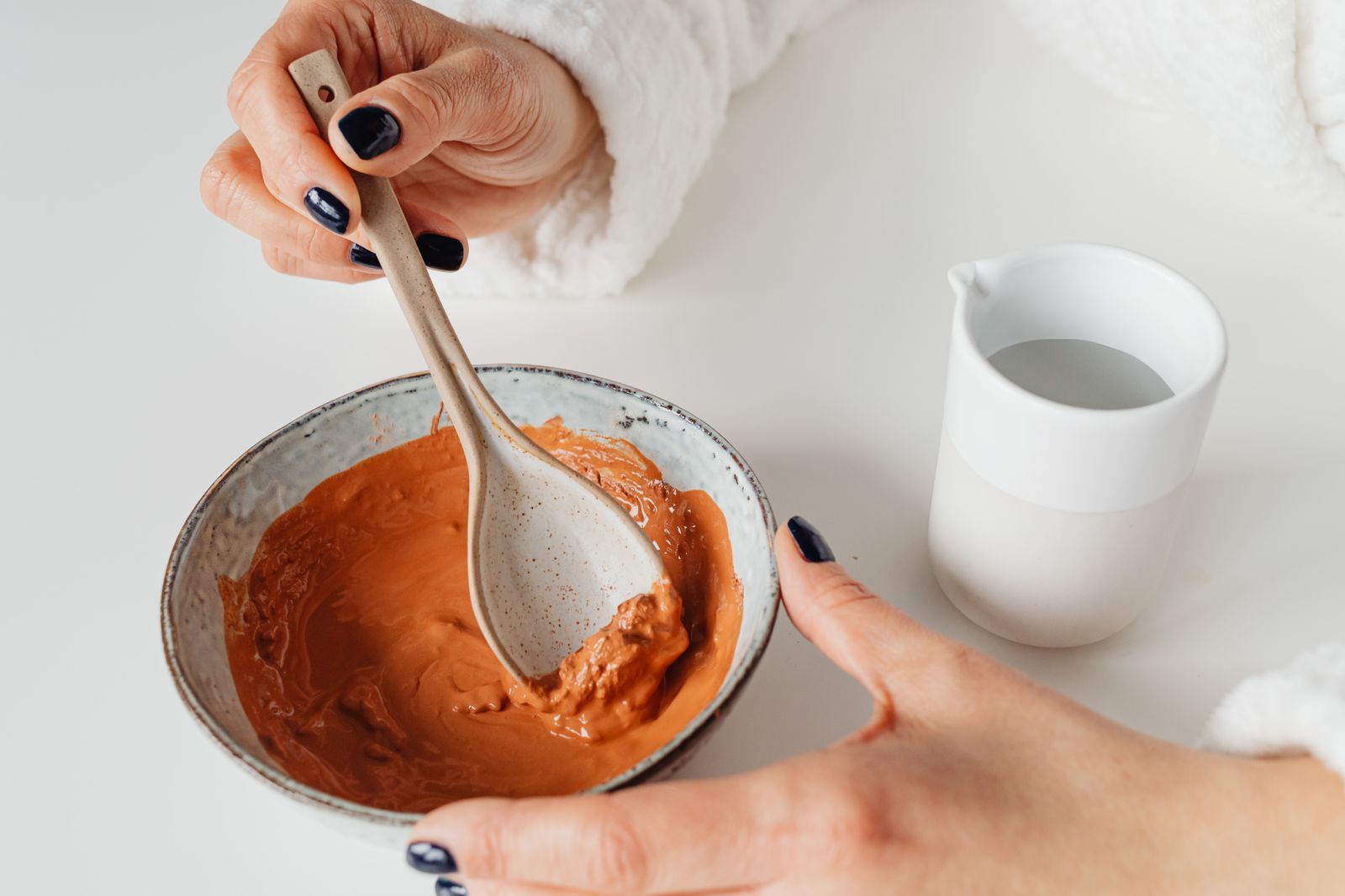 Inside the Moroccan Hammam Ritual at Kimantra: A Step-by-Step Photo Story 8 Terracotta-coloured clay paste being stirred with a ceramic spoon in a stoneware bowl, white robe sleeves and a small jug of water beside it